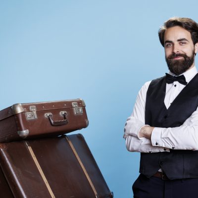 Hotel worker with baggage pile on camera, providing luxury service to guests and helping with suitcases. Professional bellhop or porter dressed in suit and tie feeling determined.